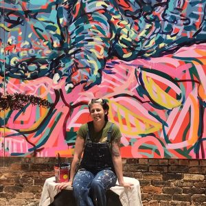 the artist of the Dinosaur of the South Mural stands in front of it with her paint supplies.